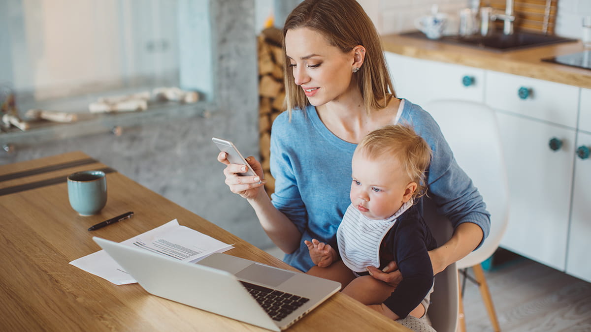 Mother at laptop looking at phone while holding baby