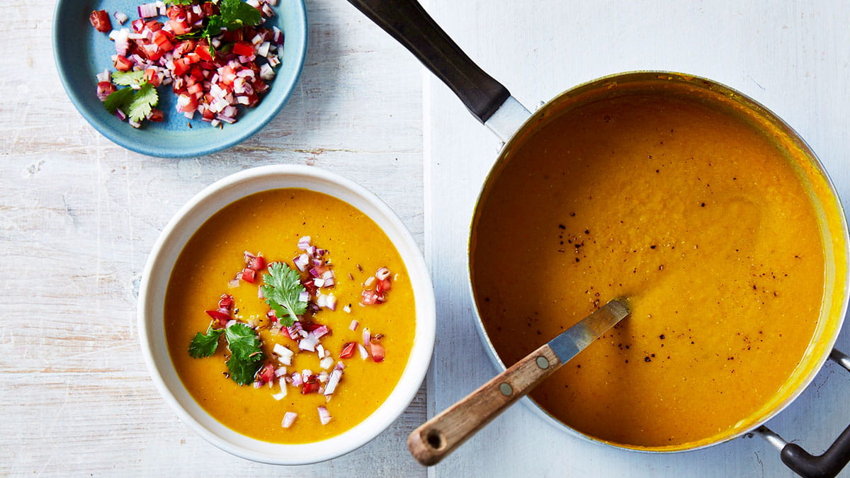 Bowl of curried pumpkin, lentil and leek soup topped with tomato, red onion and coriander, served beside a saucepan of soup.