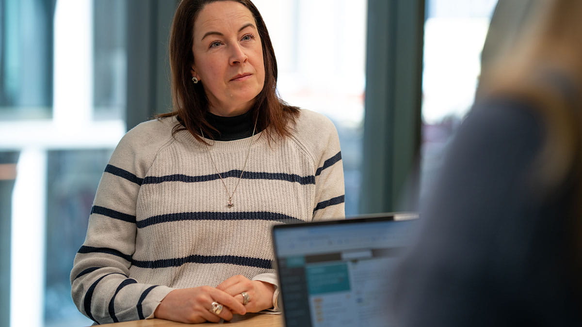 Person seated at a meeting table during a workplace conversation, with a laptop visible in the foreground