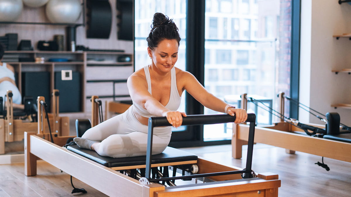Person using a Pilates reformer machine in a bright fitness studio, performing a stretching exercise with exercise equipment visible in the backgrou
