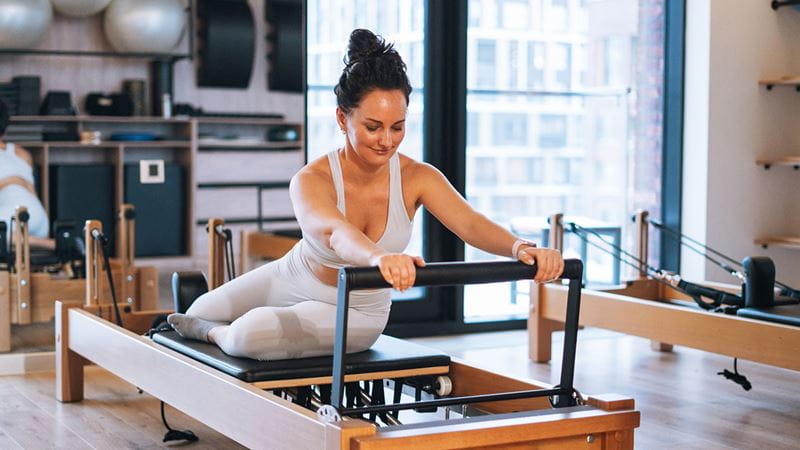 Person using a Pilates reformer machine in a bright fitness studio, performing a stretching exercise with exercise equipment visible in the backgrou