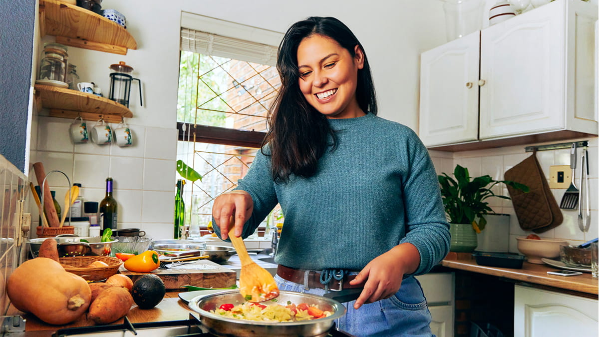 Person cooking vegetables in a frying pan on a stovetop in a home kitchen, with fresh produce including pumpkin, sweet potatoes and avocado on the bench.