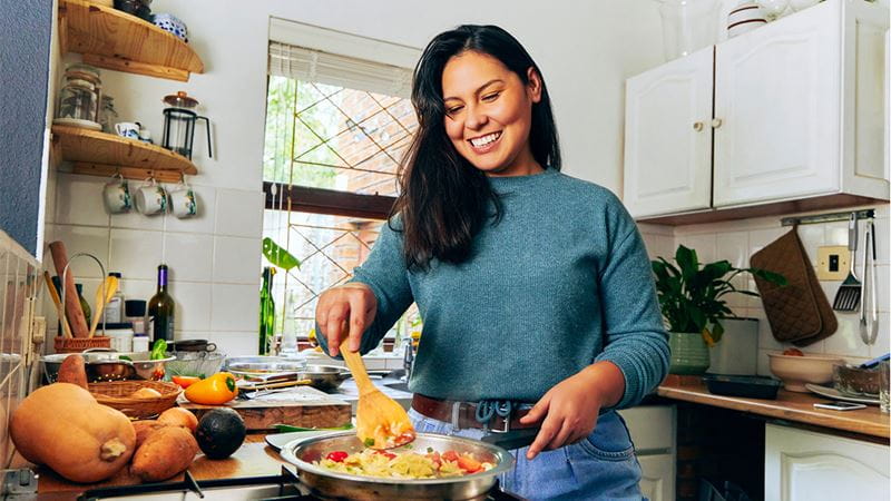 Person cooking vegetables in a frying pan on a stovetop in a home kitchen, with fresh produce including pumpkin, sweet potatoes and avocado on the bench.