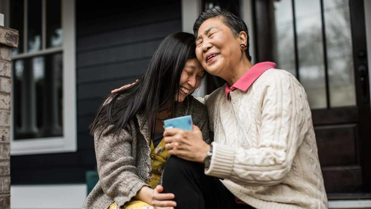 An elderly woman laughing with her adult daughter over a cup of tea outdoors