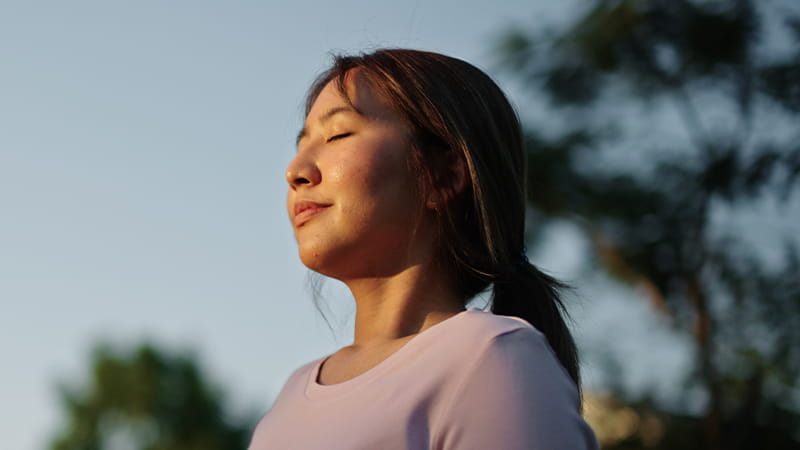 Person standing outdoors with eyes closed, taking a quiet moment for mindfulness and relaxation in a natural setting.