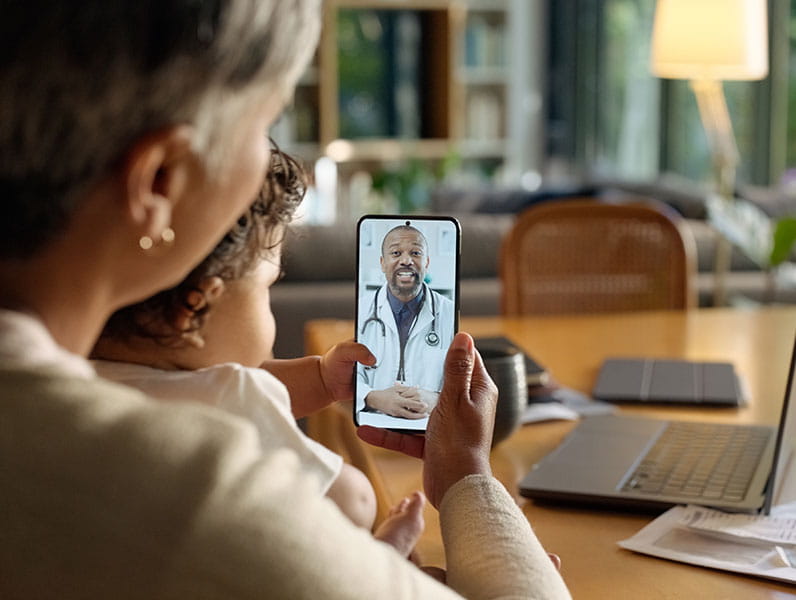 Parent holding a child while attending a telehealth consultation with a doctor on a smartphone from home