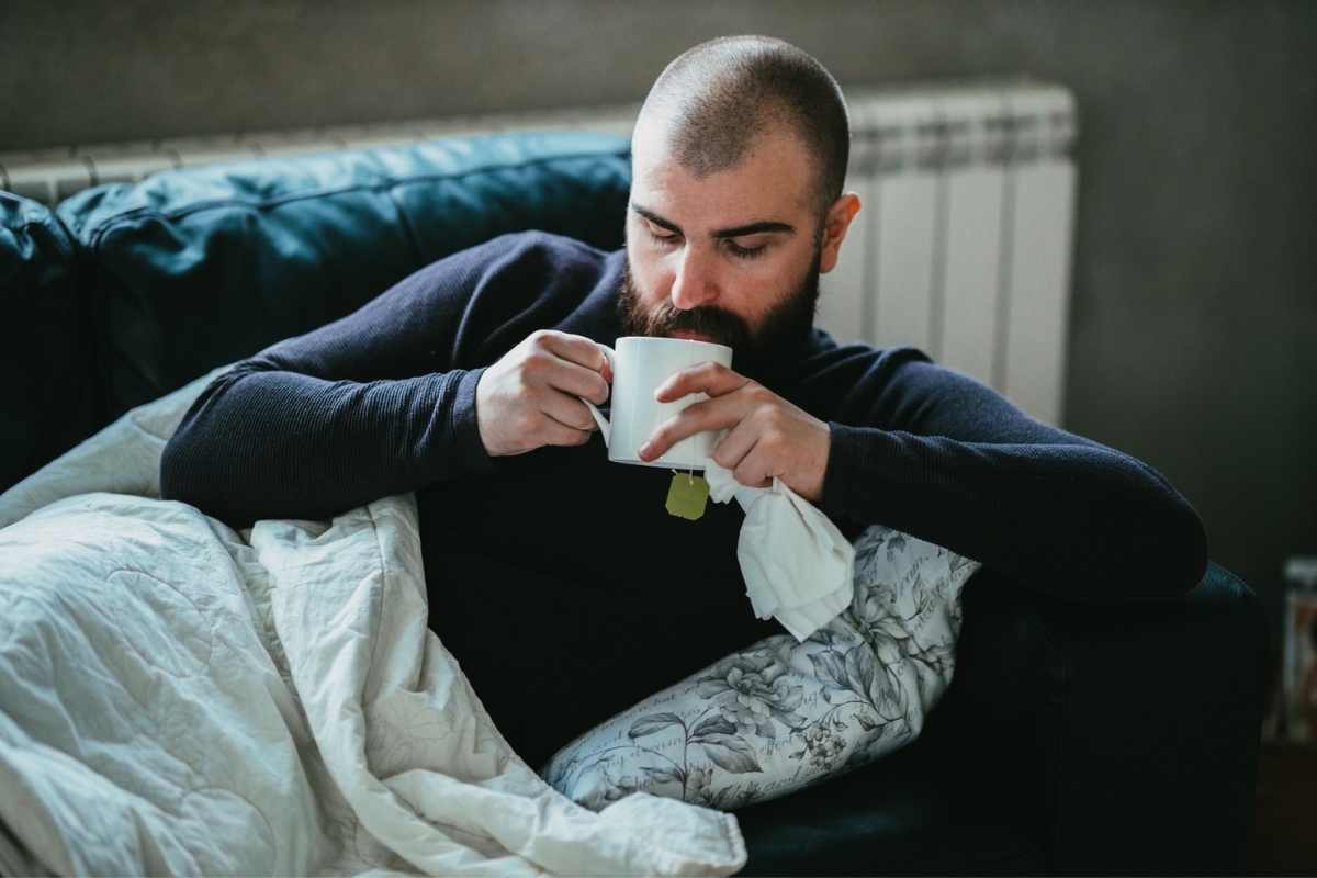 A man with mild COVID-19 symptoms, resting on the couch with a cup of tea and tissues