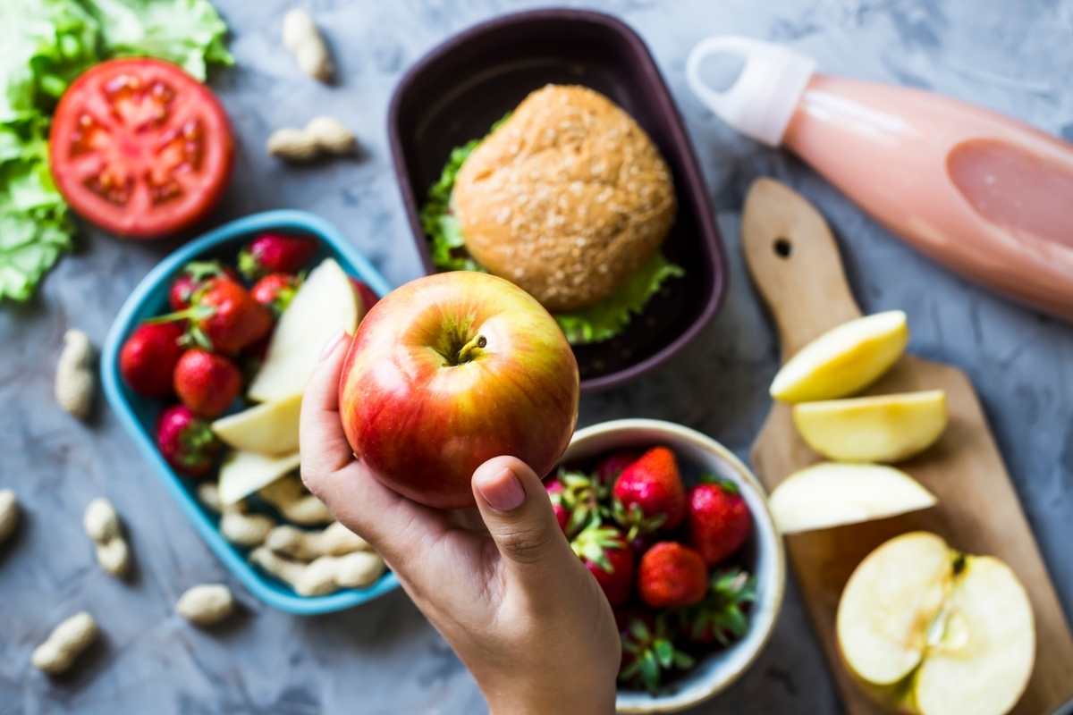 A woman's hand packing a healthy school lunchbox with fruit and a salad sandwich