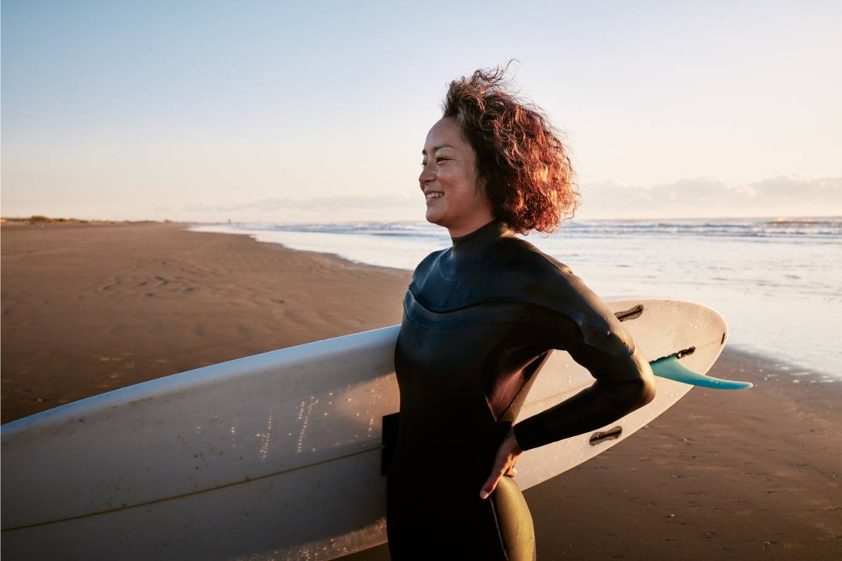 Woman stands on beach smiling with surf board