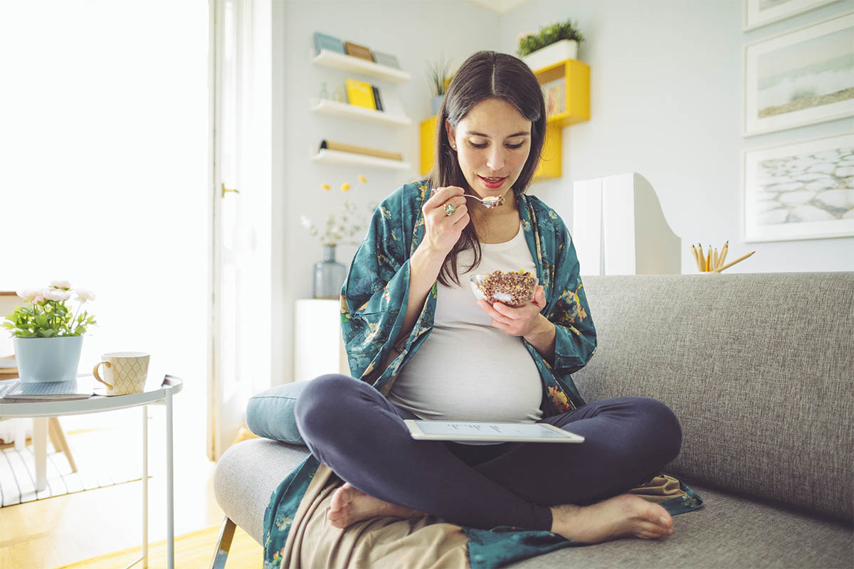 Pregnant woman eating yogurt and looking at her tablet