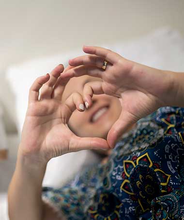 A smiling woman making a heart symbol with her hands while recovering from heart valve replacement in hospital